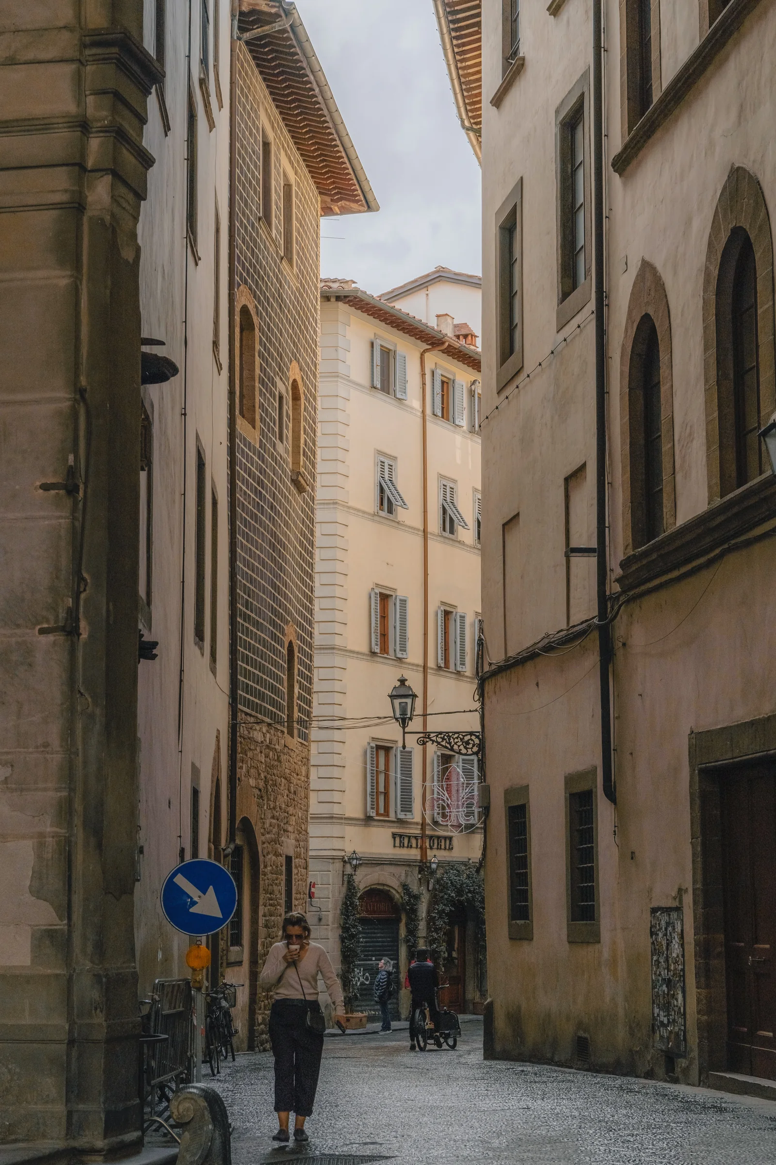 Woman walking a quiet alley between tall ochre palazzi, blue arrow sign at left