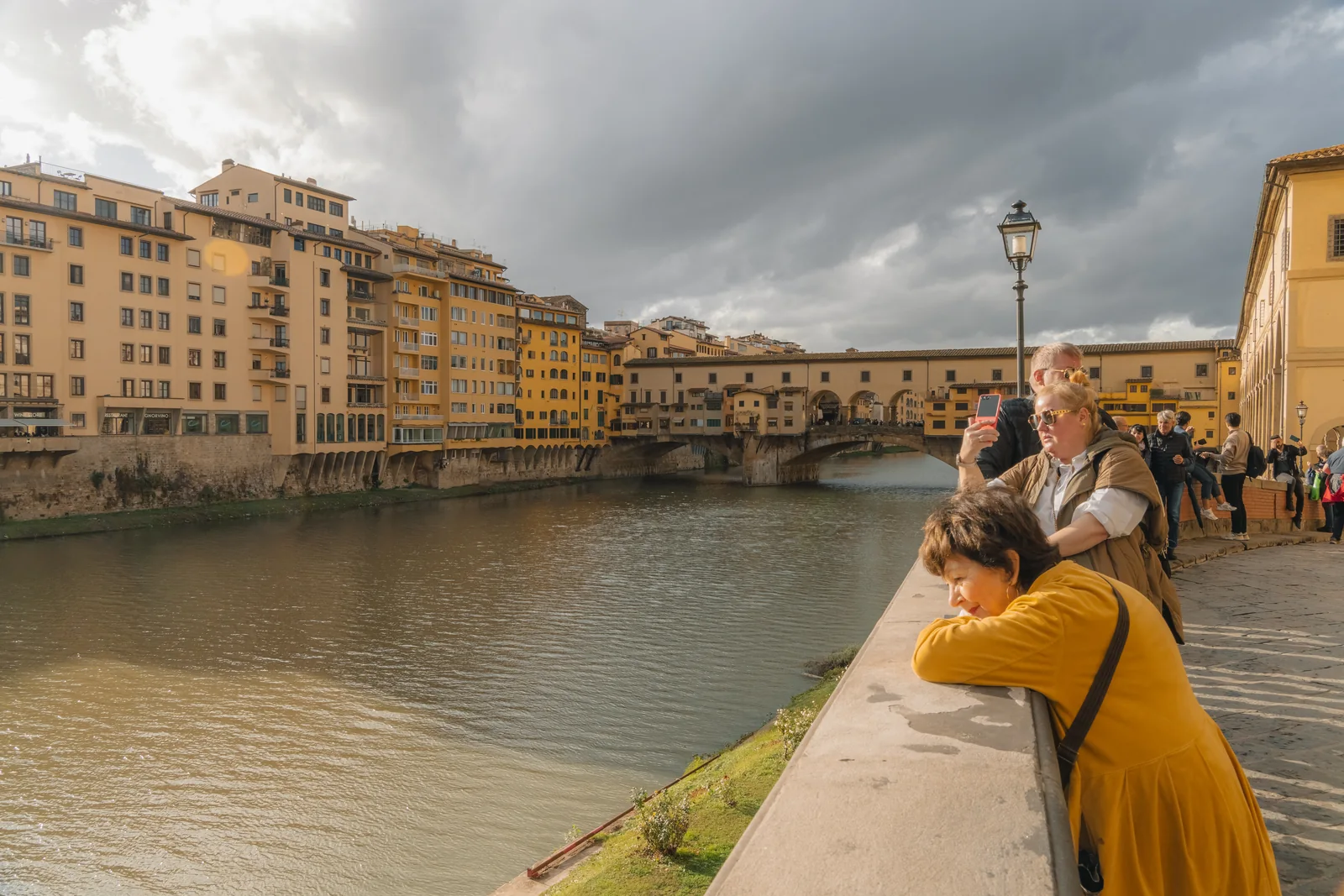 People leaning on the embankment wall watching Ponte Vecchio under heavy cloud