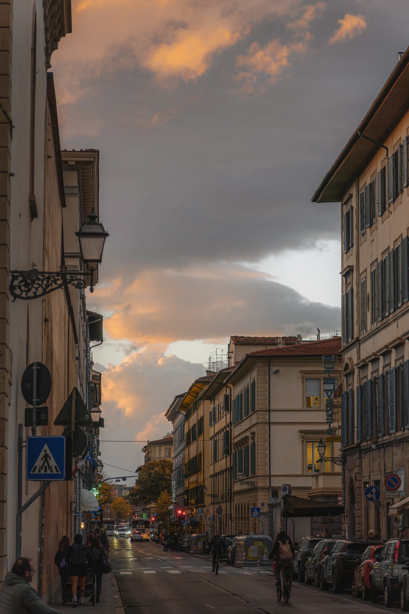 A long Florentine street at sunset, clouds catching orange light above cyclists and parked cars