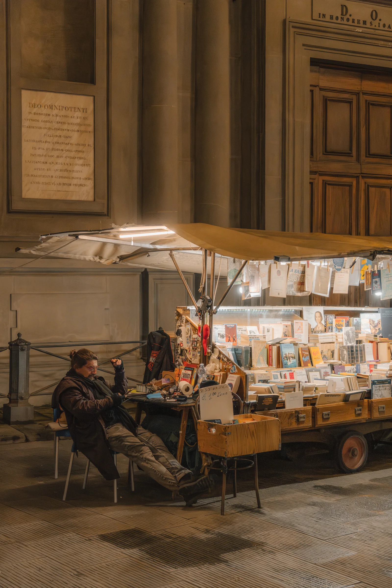 A second-hand book stall lit at night, the seller resting in a chair beside hanging newspapers
