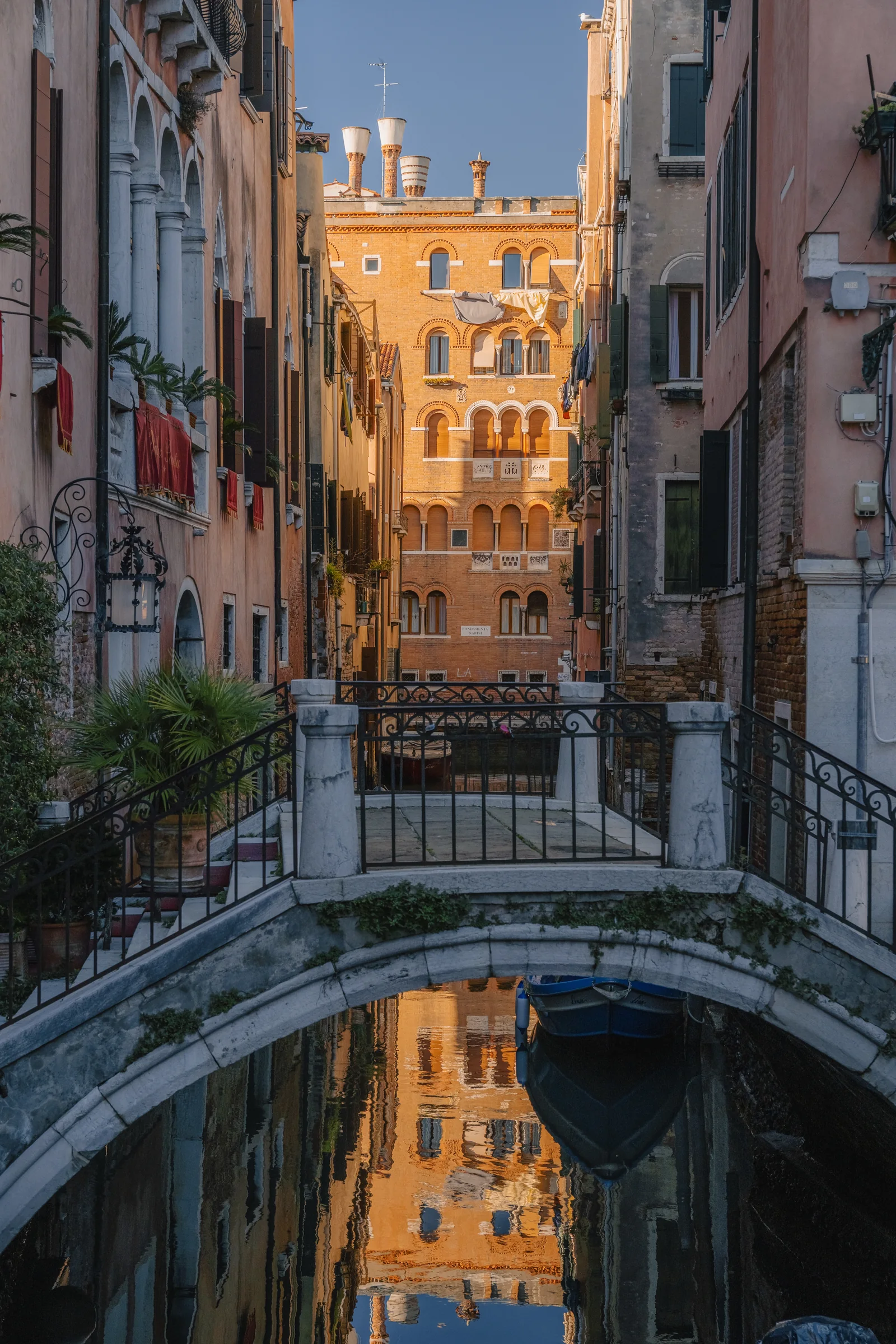 A small arched bridge over a quiet side canal, a sunlit Veneto-Byzantine facade mirrored in the water