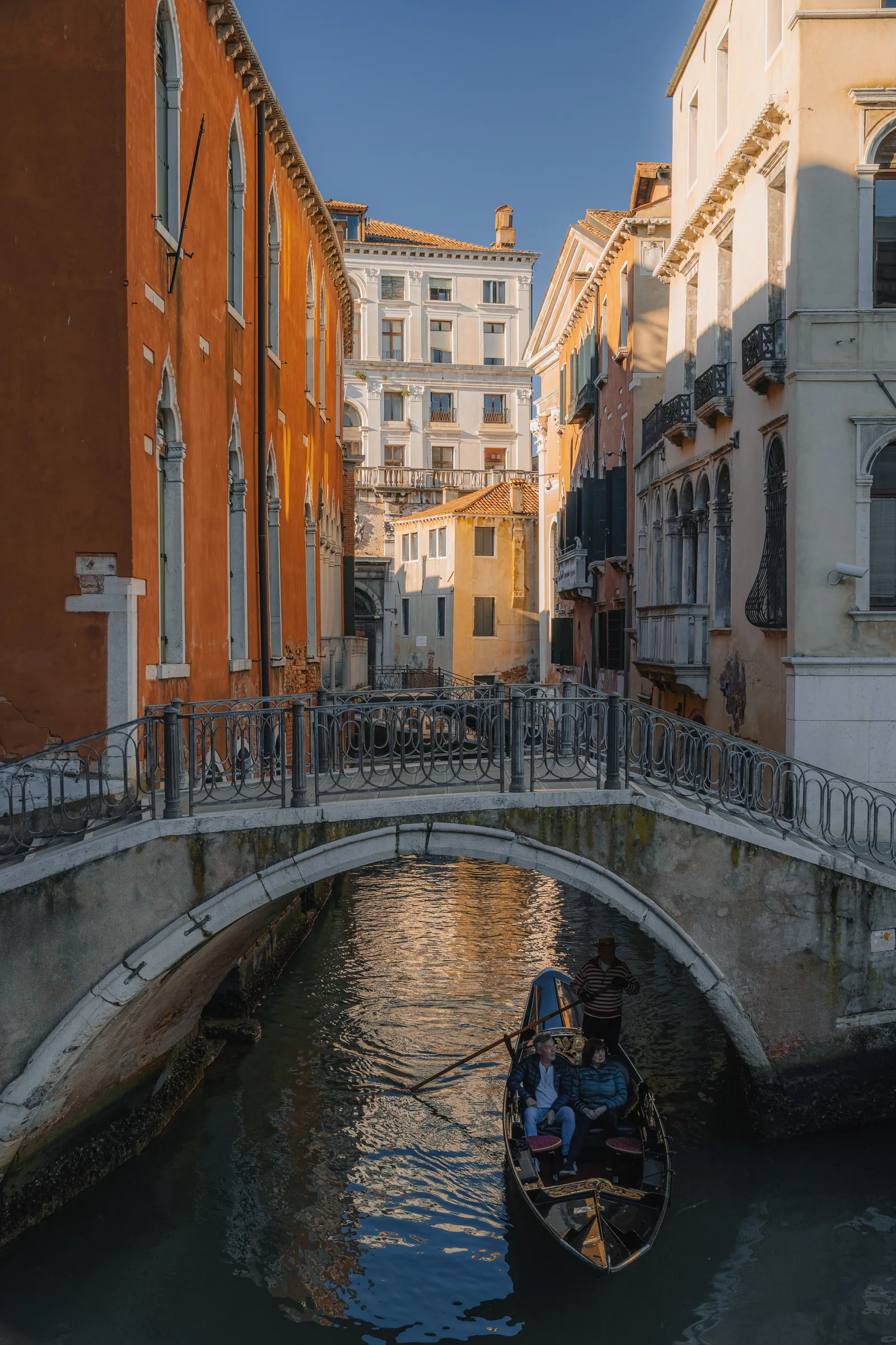 A gondola passing under a low stone bridge on a narrow canal between warm-toned houses