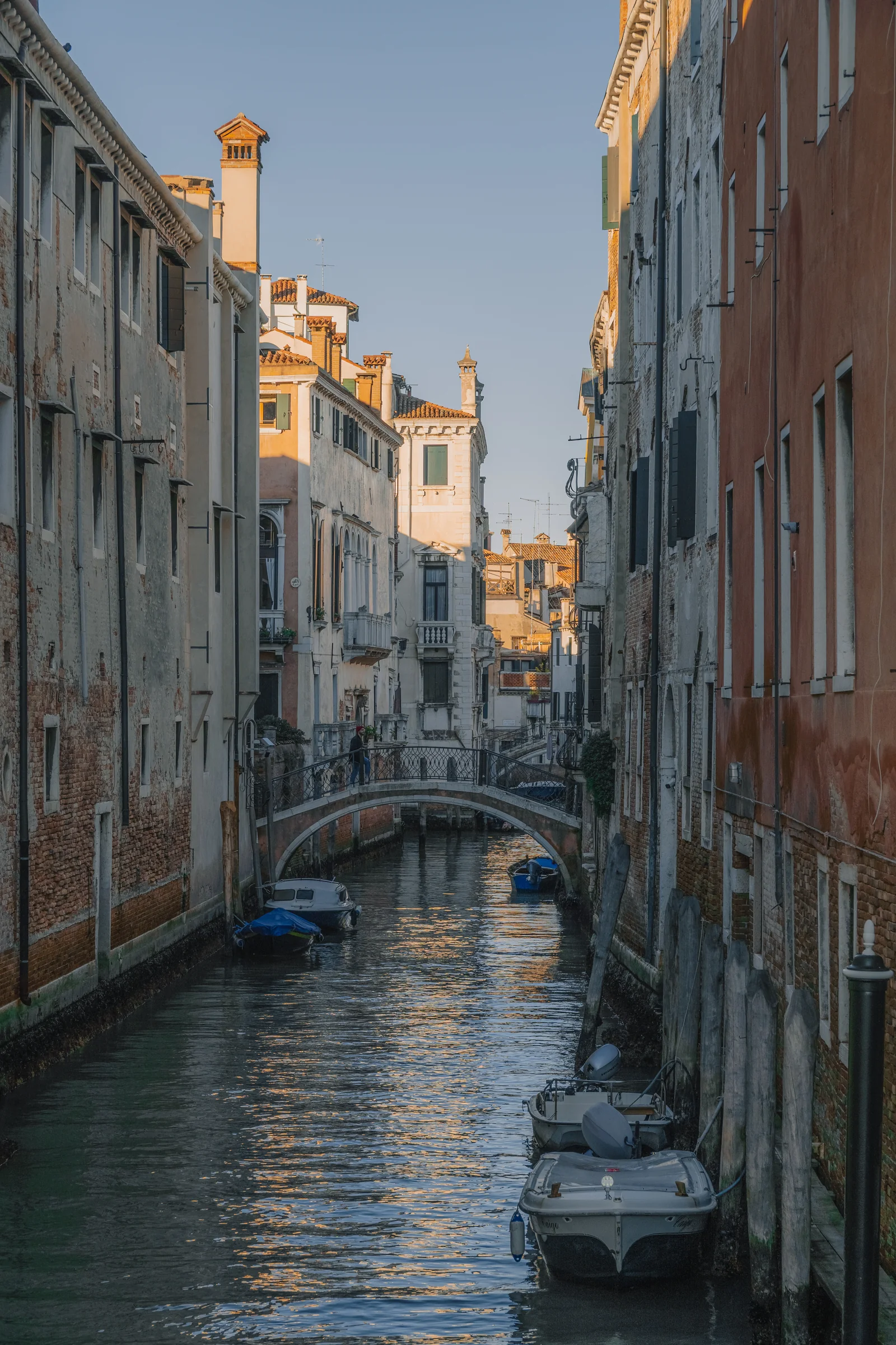 A still side canal lined with moored boats and brick facades, a footbridge catching the last light
