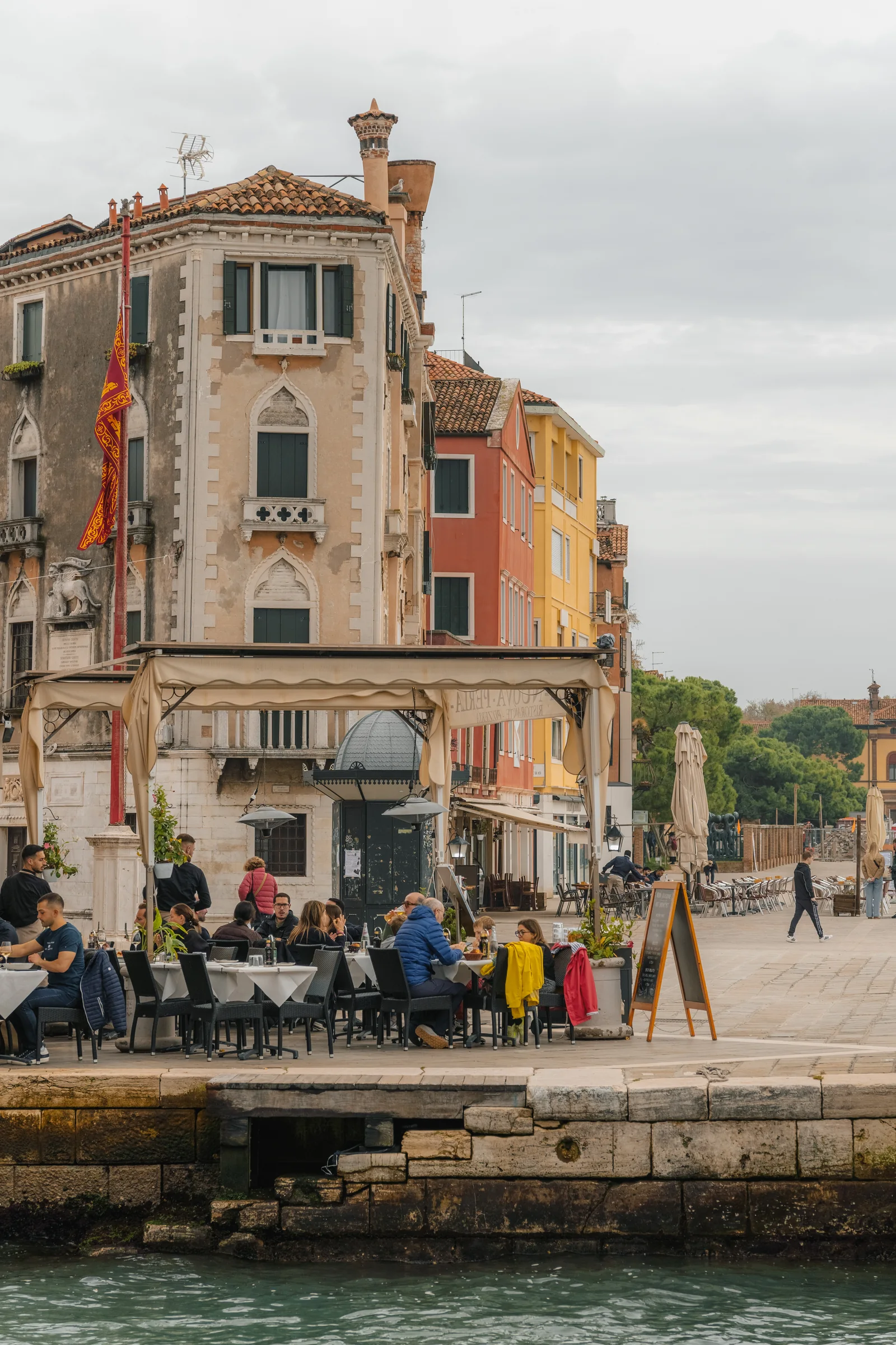 A waterside restaurant terrace busy with diners beneath a Venetian lion flag
