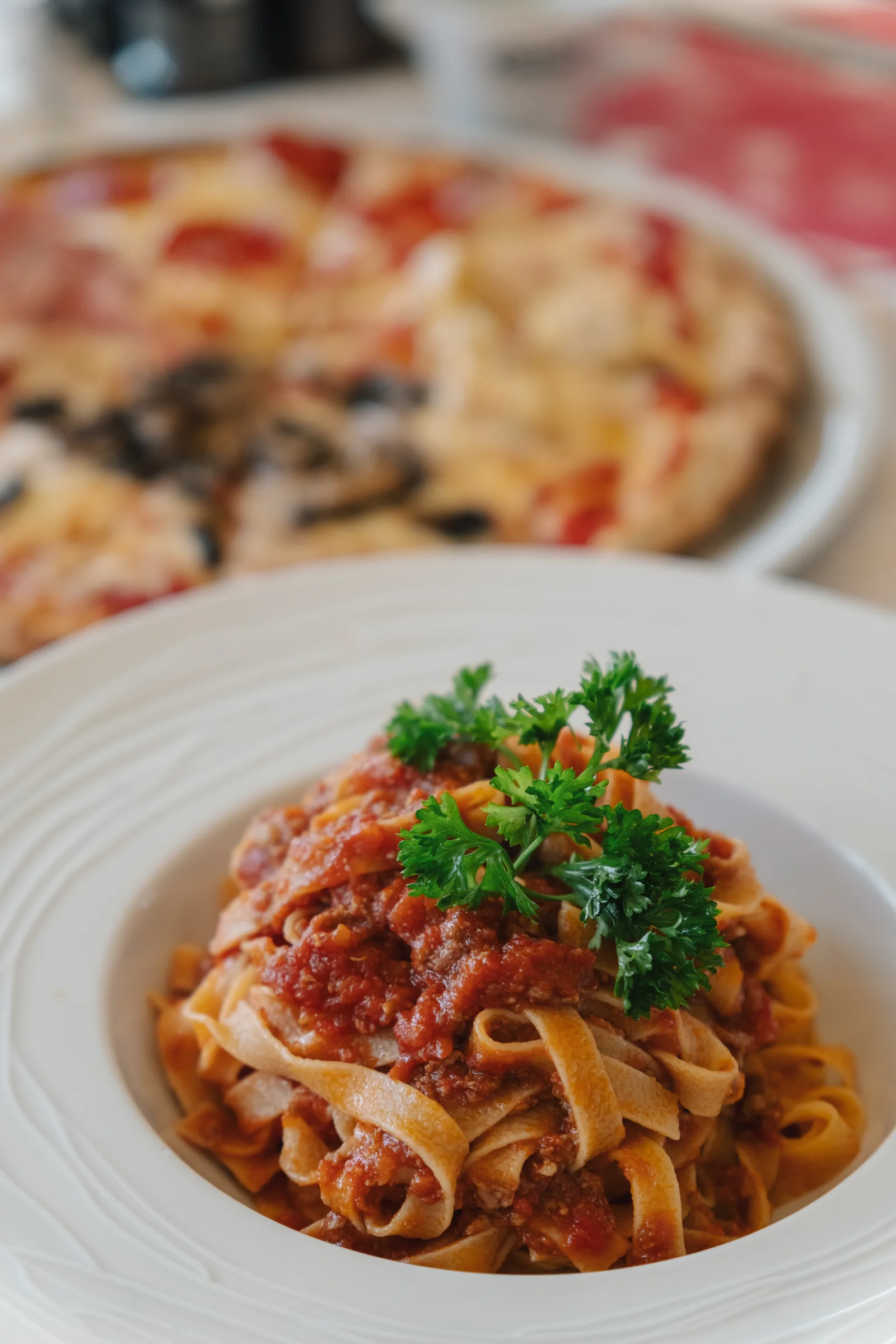 A plate of tagliatelle al ragu with parsley, a pizza waiting behind