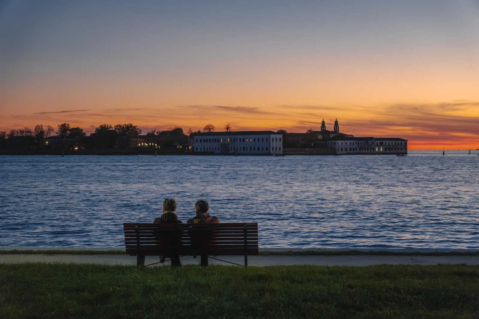 A couple on a bench watching the lagoon at sunset, an island silhouetted against orange sky