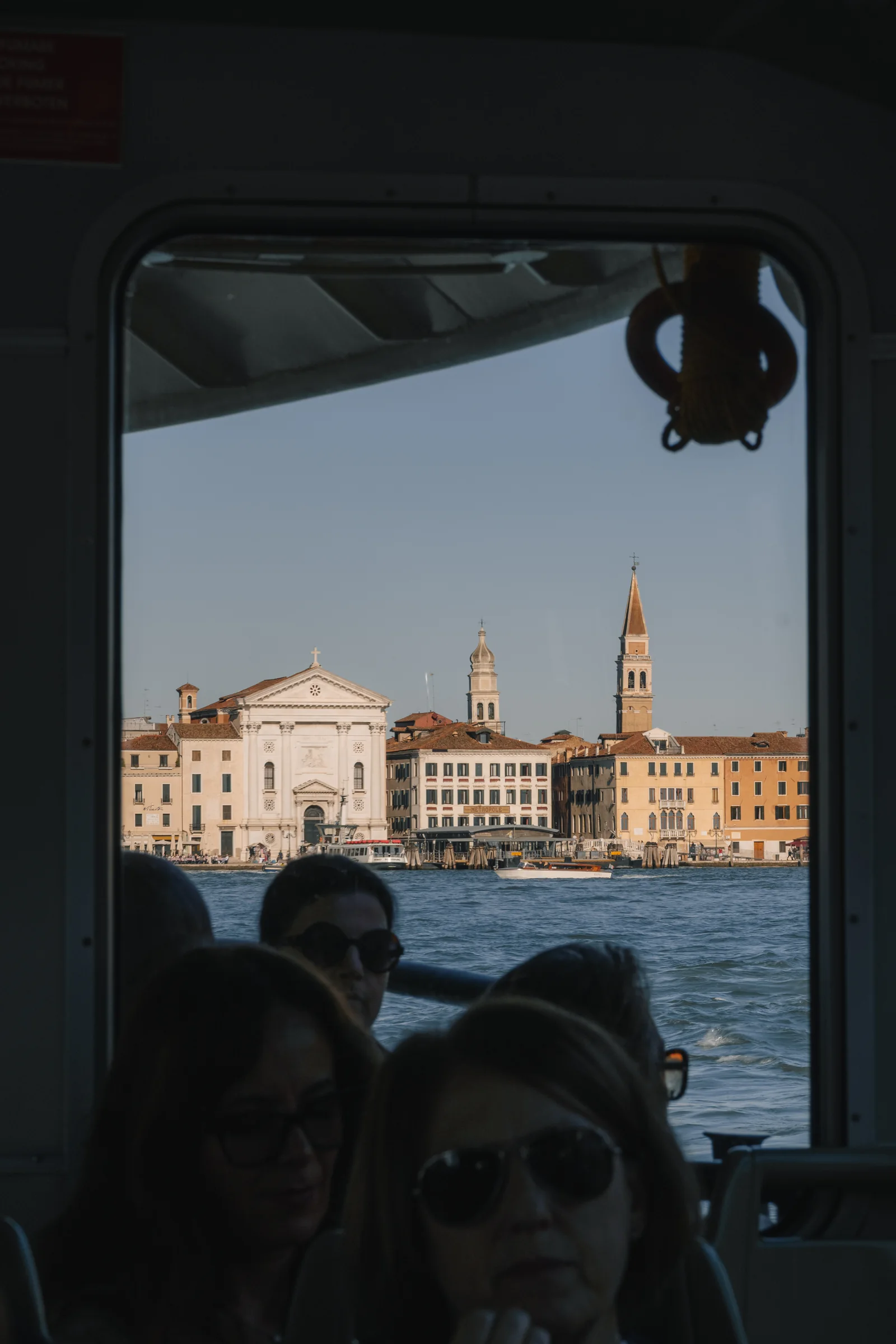 The waterfront churches and campanili seen through a vaporetto window past silhouetted passengers
